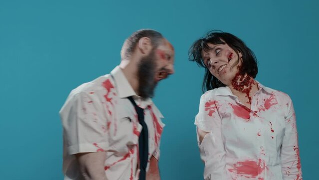 Funny spooky looking zombies biting each other and acting playful in front of camera. Crazy brain dead walking corpses with bloody and deep and wounds acting childish on blue background.