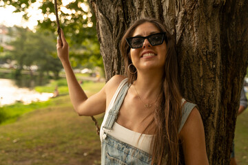 beautiful young woman with sunglasses leaning on a tree looking to camera and smiling in a park