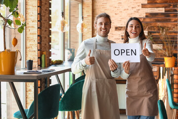 Young business owners with opening sign showing thumbs-up in their cafe