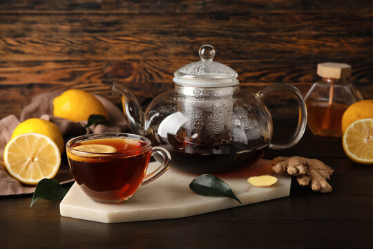 Board with glass cup of black tea, lemon, ginger and teapot on dark wooden background