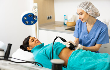 Qualified woman cosmetologist performing ultrasound cavitation body procedure for female client at aesthetic cosmetology clinic