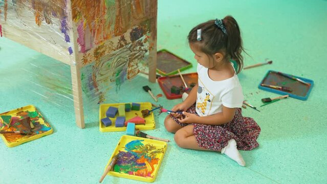 Little dark-skinned girl sitting on floor in nursery painting on plastic wrap. Creative exercise for children. Playtime. Horizontal indoor video. High quality 4k footage
