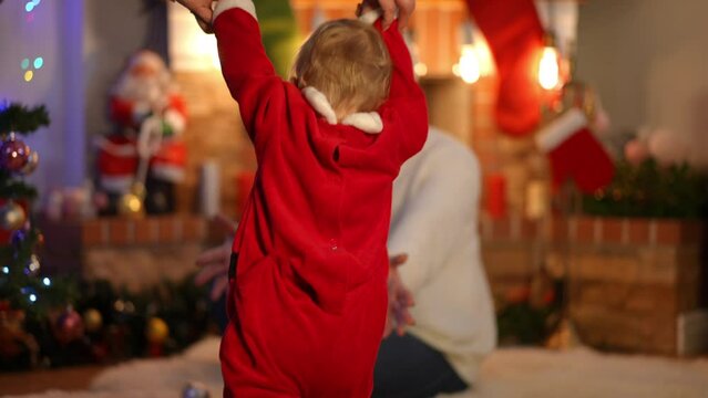 Back view little girl walking to woman sitting at Christmas tree and fireplace in living room. Caucasian toddler making steps strolling in slow motion as happy smiling mother kissing kid