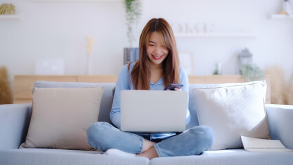 Fototapeta premium Young asian woman using computer laptop and mobile smartphone while seated on couch at home