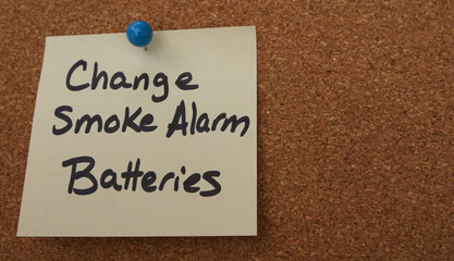 Sticky note reminder to change smoke alarm batteries, posted on a cork board.