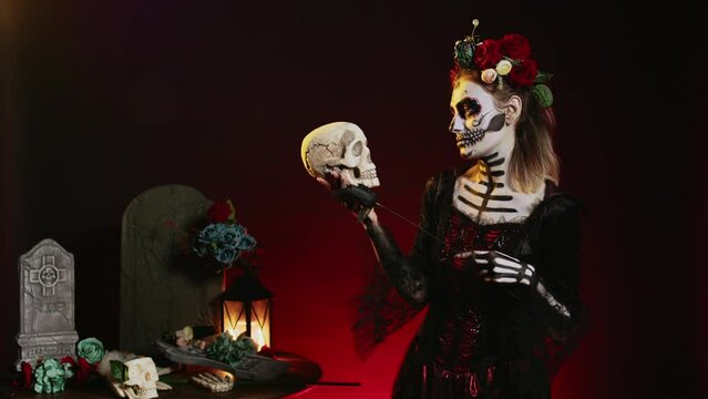 Horror scary model wearing traditional make up to celebrate mexican holiday, holding skull in studio. Looking like santa muerte or goddess of death on holy dios de los muertos celebration.