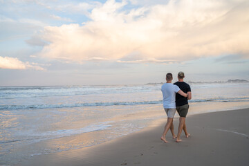 gay couple with cuddle from behind walking away on beach and fairy floss sky 