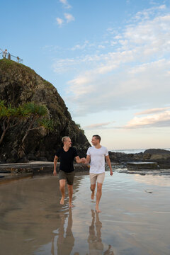 gay couple running together holding hands on beach with tree and rocks looking at each other and smiling