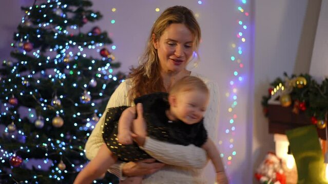 Portrait of smiling mother playing with cute little daughter in slow motion at background of Christmas lights at home. Happy Caucasian woman enjoying New Year's eve with toddler child indoors