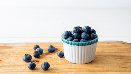 close view of a small white ceramic bowl filled with fresh blueberries. Placed on top of a wooden table