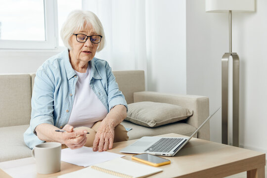 An Attentive, Sensible Elderly Woman Is Sitting At Home Working Remotely And Looking At A Piece Of Paper That She Holds In Her Hand