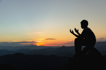Silhouette of a young man praying to God on the mountain at sunset background. Woman raising his hands in worship. Christian Religion concept.