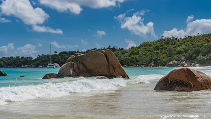 Picturesque granite boulders in the surf on the beach of a tropical island. The waves beat against the rocks, foam and spread on the sand. Splashes in the air. A green hill against a blue sky clouds