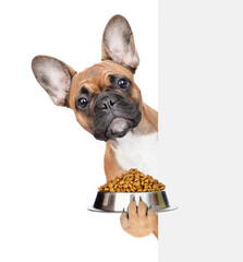 Funny french bulldog puppy holding bowl of dogs food behind empty white banner. isolated on white...