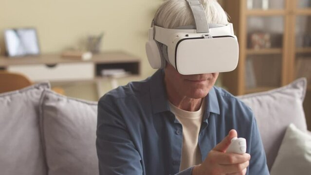 Grey haired senior man in vr headset using vr controller while playing dynamic vr game on sofa in living room