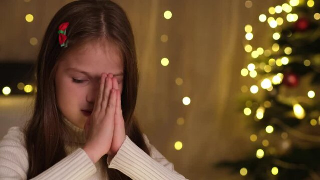 Kid girl is praying in decorated room on background of Christmas tree and glittering lights. Child pray for something good for Christmas. Holidays, childhood, children dreams and family concept 