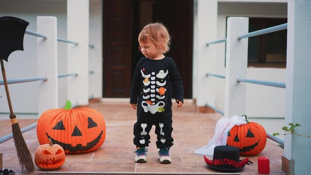Sweet unhappy boy in skeleton costume celebrate Halloween alone. Sad lonely toddler at home at background of decorated with pumpkin lanern house. Trouble concept