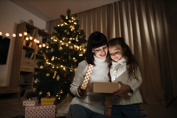 Merry Christmas. Family of two mother and little cute child girl with magic gift at home near Christmas tree. Happy mom and cheerful daughter unboxing fabulous gift.