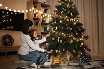 Happy small preschool caucasian child girl decorating Christmas tree with happy young mother, putting toys on branches, enjoying preparing for New Year celebration at home, miracle time concept.