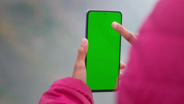 Closeup shot of Indian girl wearing jacket and using mobile phone with Green screen. Green screen mockup template of Mobile phone, Chroma key.  Finger swiping up in green screen of the mobile