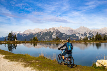 Mountainbiker macht eine Pause am Kaltwassersee in Seefeld/Tirol. Der schneebedeckte Gipfel am linken Bildrand ist die Zugspitze - obrazy, fototapety, plakaty