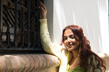 Beautiful young woman with long curly hair and wearing an elegant green dress. The woman is standing by a window holding on to the grille of the old building in Seville. Holiday concept.