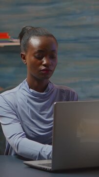 Vertical video: African-american woman looking at camera smiling working at computer from home lare at night sitting at desk in living room. Black businesswoman typing on computer in evening.