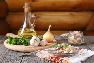 Pickled mushrooms near vegetables and herbs are on a wooden table with a log wall background. 