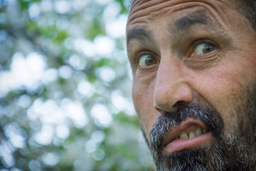 An adult gray-haired bearded man grimaces in nature. Close-up