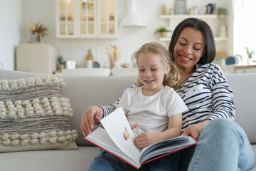 Happy foster mother, preschool adopted daughter reading fairytale book, sitting on couch at home