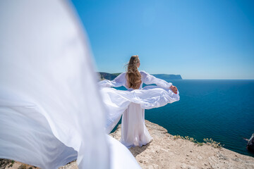 Fototapeta na wymiar Blonde with long hair on a sunny seashore in a white flowing dress, rear view, silk fabric waving in the wind. Against the backdrop of the blue sky and mountains on the seashore.