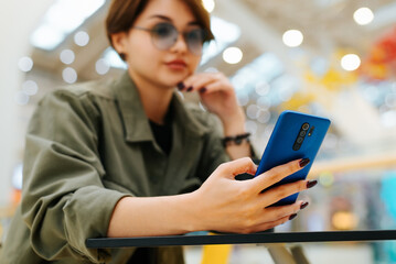 Asian young woman using smartphone while sitting in cafe in food court of mall. Close-up of stylish woman in glasses holding phone while sitting at table, selective focus on mobile phone