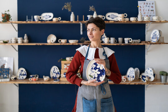 Craftswoman posing with handmade plate after decorating pottery with hand drawn ornament in ceramics studio or workshop. Female ceramist creating handmade kitchenware on classes