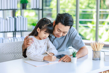 Millennial Asian lovely happy family father helping teaching young little girl daughter using touchscreen tablet computer and notebook writing doing homework on working desk in living room at home
