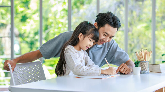 Millennial Asian lovely happy family father helping teaching young little girl daughter using touchscreen tablet computer and notebook writing doing homework on working desk in living room at home
