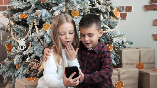 Cute happy cheerful kids brother and sister having online video call with parents. Children laugh waving hands congratulate grandmother on Christmas. Brother hugs sister near the Christmas tree.