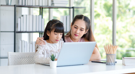 Millennial Asian happy family mother smiling helping supporting teaching little girl kid daughter studying learning doing online school homework via laptop notebook computer in living room at home