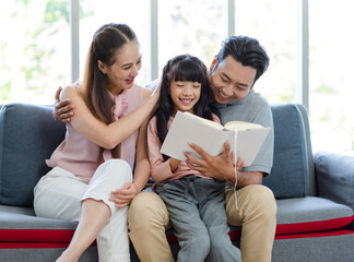 Millennial Asian happy family father mother sitting on cozy sofa couch smiling together teaching little girl kid daughter learning studying reading fiction novel textbook in living room at home