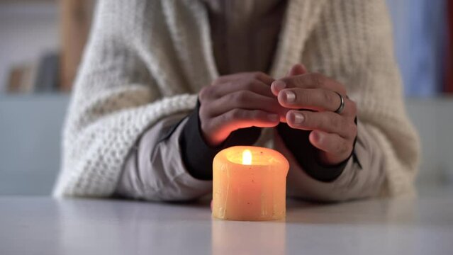 man dressed in a warm winter clothes sits at home at a table and warms his hands from a burning candle, close up. Concept of no heating in winter at home.