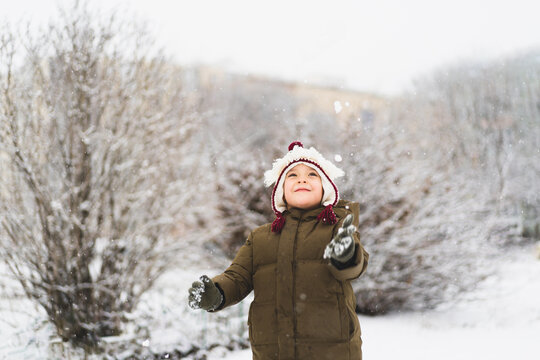 Cute Little Boy In Funny Winter Hat Walks During A Snowfall. Outdoors Winter Activities For Kids.