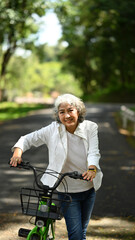 Smiling middle age woman cycling in summer park on beautiful day. Retirement people lifestyle, outdoor activity concept