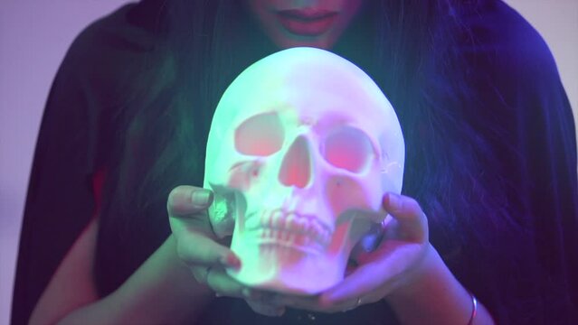 Halloween Witch woman holding human skull lantern in her hands. Beautiful young woman doing witchcraft at night. Darkness. Scary scene. Over spooky dark blue magic background 