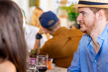 Engaged couple in a bar drinking wine. Red-haired man with beach hat in foreground.