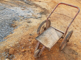 Wheelbarrow on construction area, Metal trolley with three wheels, Rusty old cart on the background of piles of stones and sand.