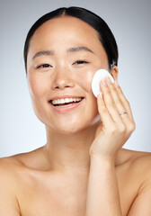 Skincare, beauty and woman with cotton pad for facial cleaning against a grey studio background. Happy, smile and portrait of a young Asian model with cotton for face wellness, clean body and makeup © Kirsten D/peopleimages.com