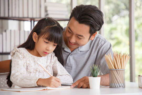 Children education and home school concept : Young asisn father pleased to see little daughters' study success. Excited smiling small child girl enjoying learning and writing with pleasant dad at home