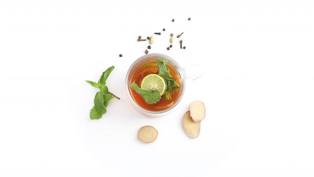 lemon tea in a transparent glass cup with sliced lemon and ginger mint leaves cloves cardamom black pepper in white background rotating top shot
