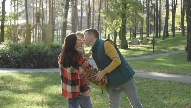 A man kisses a woman who holds a child in her arms, and throws up fallen tree branches. Family in the autumn park. Outdoor recreation. Happy family outdoors. High quality 4k footage