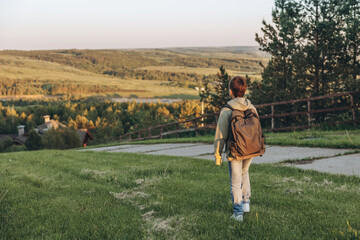 Tourist with backpack walking on top of hill in grass field and enjoying beautiful landscape view. Rear view of teenage boy hiker resting in nature. Active lifestyle. Concept of local travel