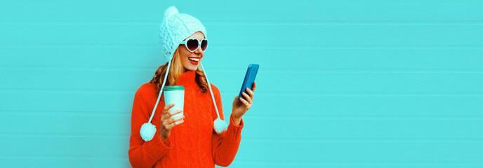 Winter portrait of happy smiling young woman with smartphone and coffee cup wearing red knitted sweater, white hat with pom pom, heart shaped sunglasses on blue background